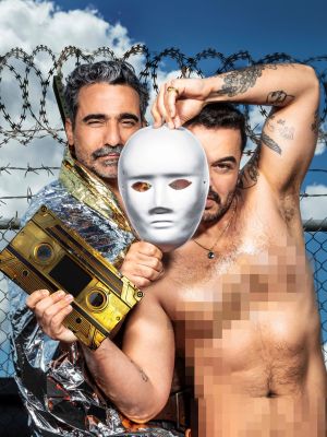 Auf dem Foto sieht man zwei Schauspieler vor einem Stacheldrahtzaun stehen. Der vordere Schauspieler ist oberkörperfrei und hält eine goldene Kassette in seiner rechten Hand. Seine Brust ist verpixelt. Seinen linken Arm hebt er über seinen Kopf und in der Hand hält er eine weiße Maske, die sein Gesicht halb verdeckt. Der hintere Schauspieler hält den unteren Teil derselben Maske mit seiner rechten Hand fest. Er ist in eine silberne Rettungsdecke gewickelt.